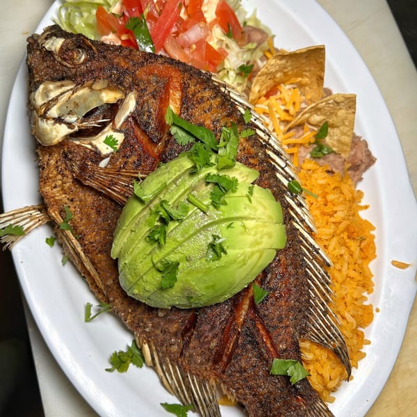 A whole fried fish topped with avocado slices and cilantro, served with rice, beans, and salad.