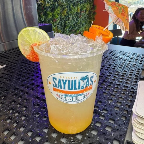 Sayulitas branded drink with lime, orange peel, and a cocktail umbrella on a black grate.