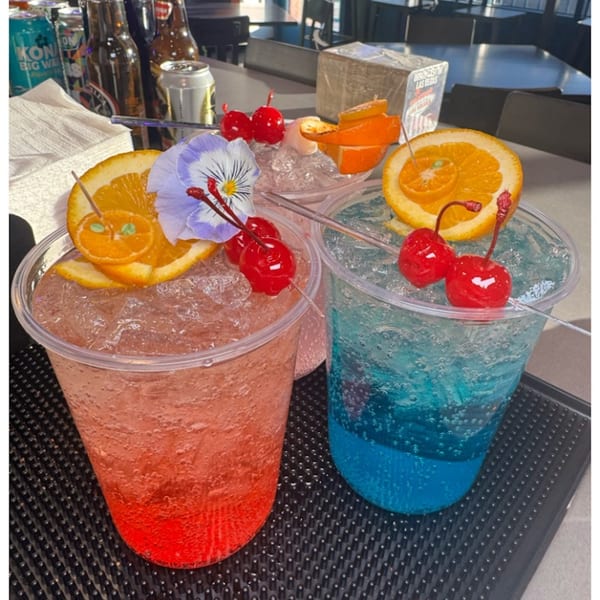 Two vibrant, colorful drinks with fruit and flower garnishes, on a bar mat with bottles in the background.