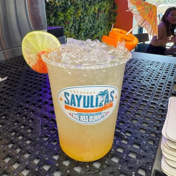 Sayulitas branded drink with ice, lime, orange slices, and a small umbrella on a dark textured surface.