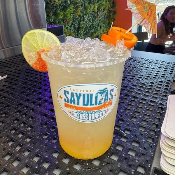 A Sayulitas branded drink with ice, a lime slice, orange peels, and a small umbrella.