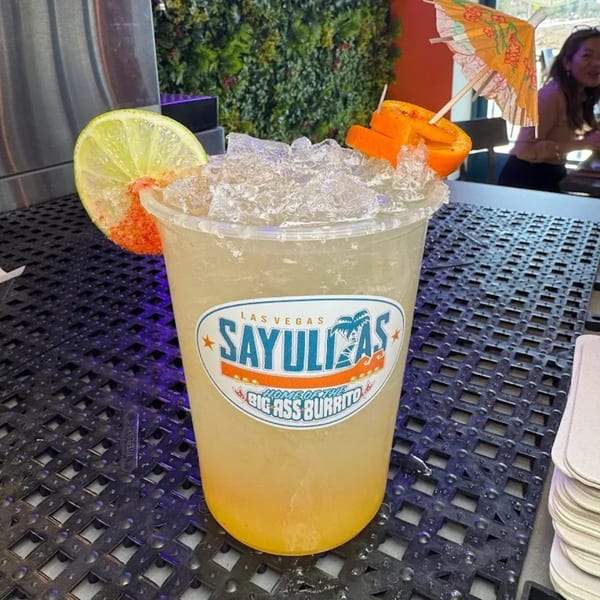 Sayulitas branded margarita with lime, orange, and a small umbrella on a dark textured surface.