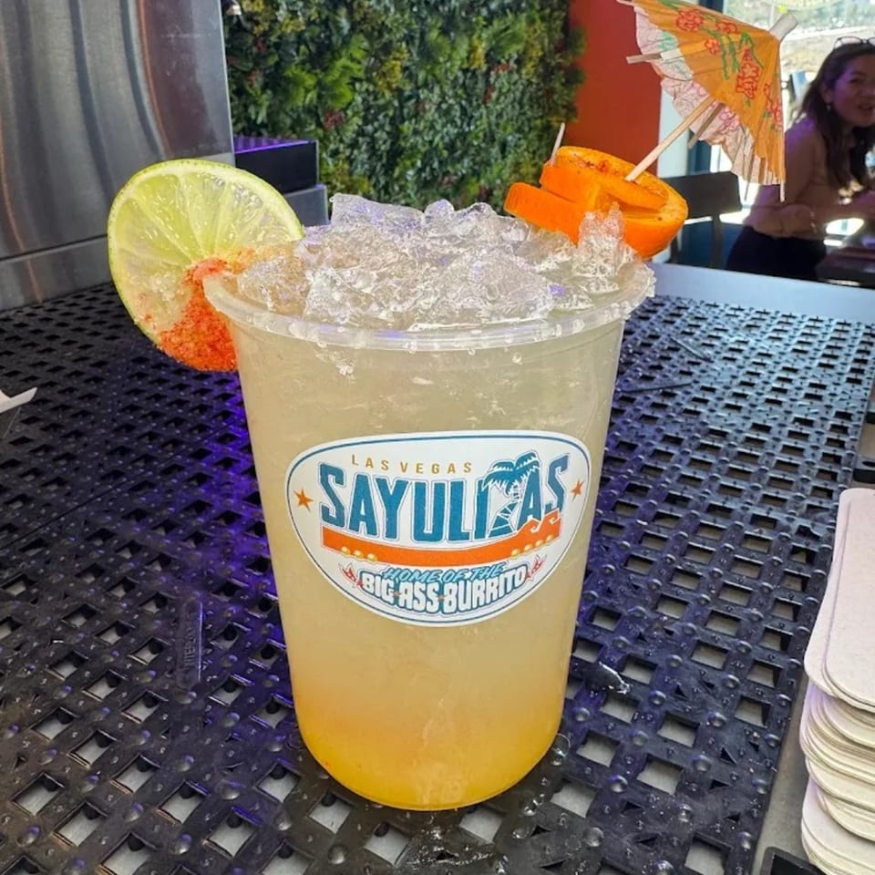 Sayulitas branded margarita with lime, orange, and a small umbrella on a dark textured surface.