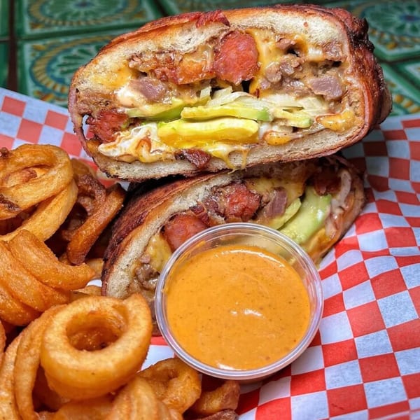 Sayulitas' Mexican torta sandwich, cut in half, with curly fries and dipping sauce on checkered paper.