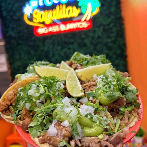 Sayulitas' street tacos with meat, cilantro, onions, avocado salsa, and lime wedges under a neon sign.