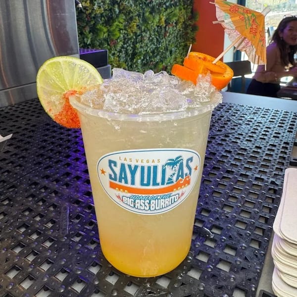 Sayulitas branded drink with ice, lime, orange slices, and a small umbrella on a dark textured surface.