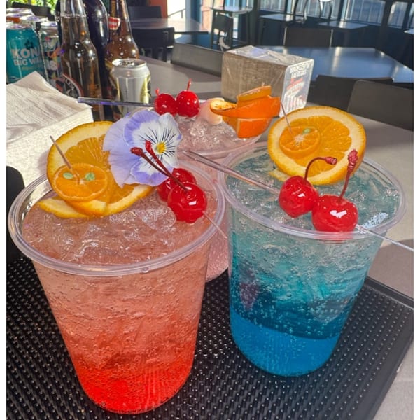 Three vibrant Sayulitas cocktails, garnished with oranges, cherries, and a purple flower, on a bar.