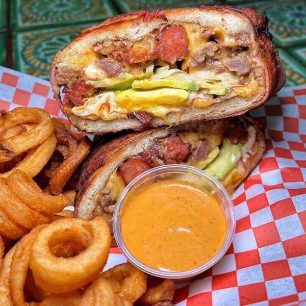 A Sayulitas Mexican torta sandwich, cut in half, with curly fries and a side of dipping sauce.