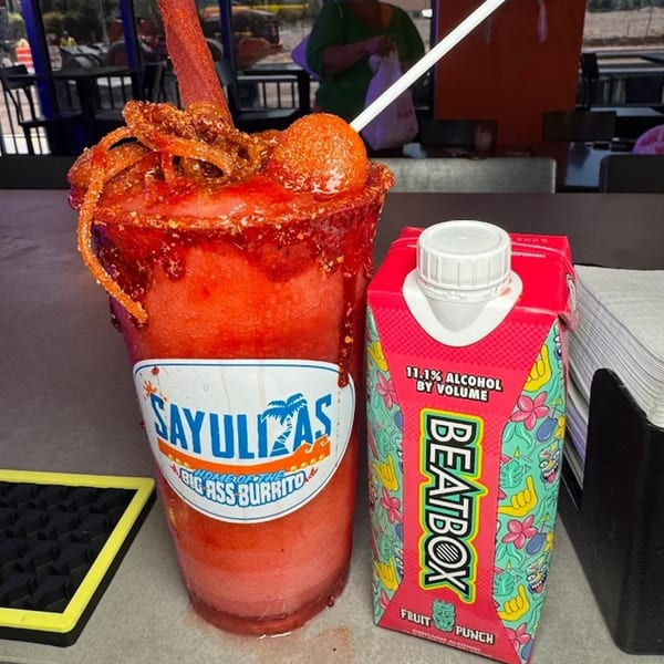 A Sayulitas chamoy-rimmed drink with a tamarind straw next to a BeatBox Fruit Punch carton.