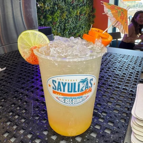 Sayulitas branded margarita with lime, orange slices, and a cocktail umbrella on a dark textured surface.